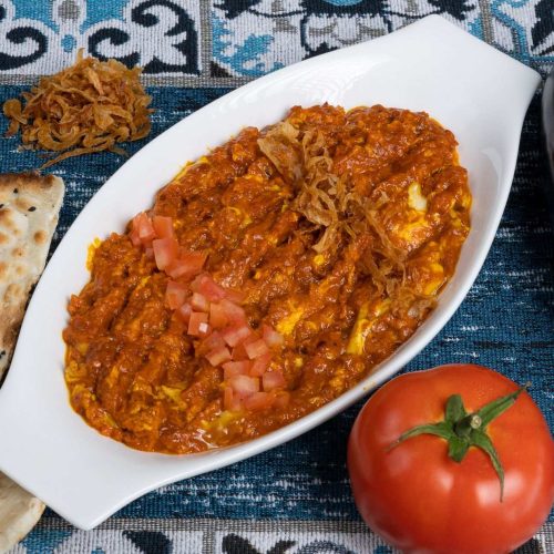 Cooked eggplant and tomato dip in a white serving dish topped with diced tomato and fried onions, with flatbread and a fresh tomato